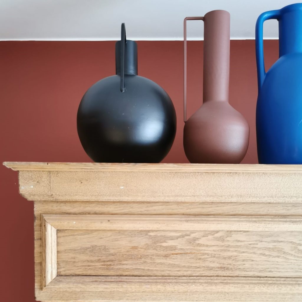 3 vases de couleur noire, terracotta et bleu roi sur un meuble en bois devant un mur terracotta.