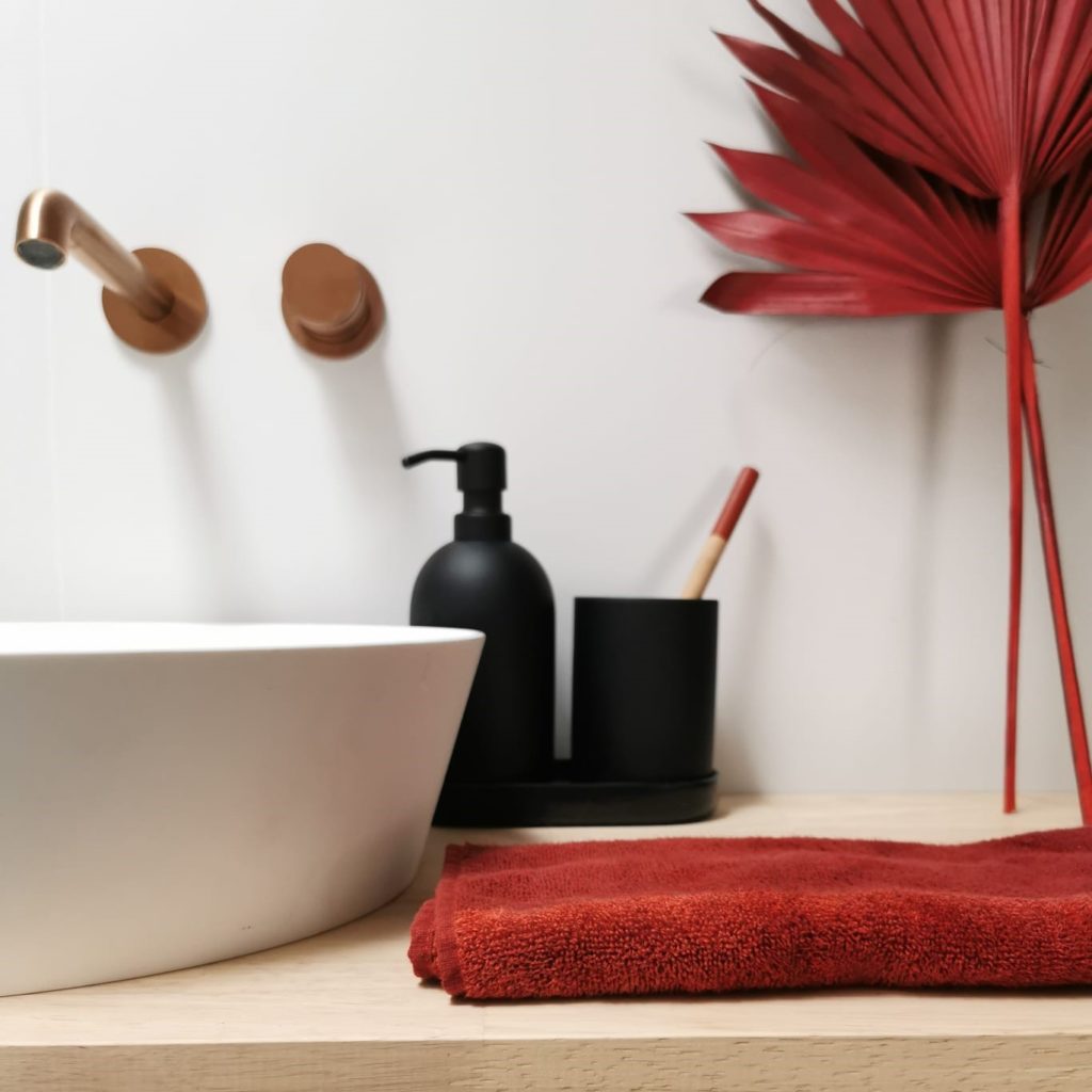 Meuble de salle de bain en bois avec vasque blanche. Essuie-main et décoration posés dessus de couleur rouge.